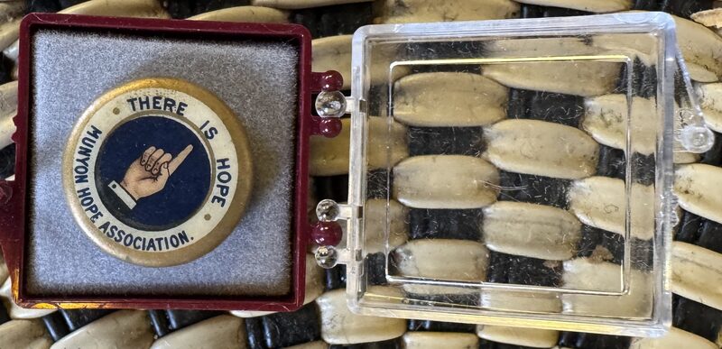 A round pin with a blue center showing an illustration of a pointing hand and the text 'THERE IS HOPE' and 'UNION HOPE ASSOCIATION' is set in a square red display case lined with gray felt. To the right sits a clear rectangular plastic box lid on a background of alternating cream-colored elongated beads threaded on black cords.