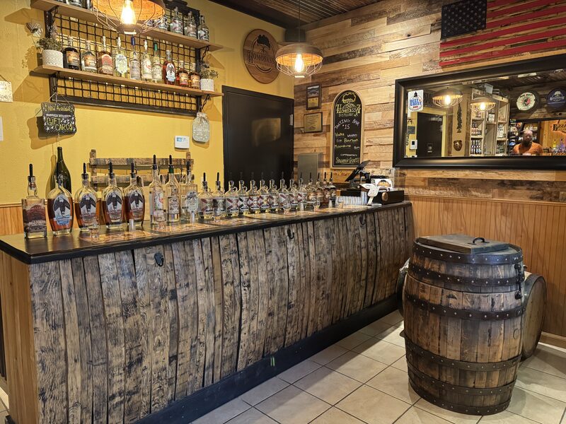 Interior of a rustic tasting room with a long wooden counter faced with barrel staves displaying numerous bottles of spirits with pour spouts, shelves of liquor and a chalkboard menu on yellow and wood-paneled walls. Pendant lights hang above, an American flag and a large mirror reflect the room (including a person), and a wooden barrel serves as a side table on a tiled floor.