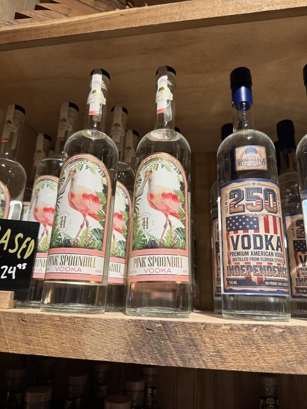Clear glass vodka bottles on a wooden shelf, including three "Pink Spoonbill Vodka" bottles with labels depicting a pink spoonbill bird and tropical foliage, and a "250 Vodka" bottle with a red-white-blue American flag-style label.