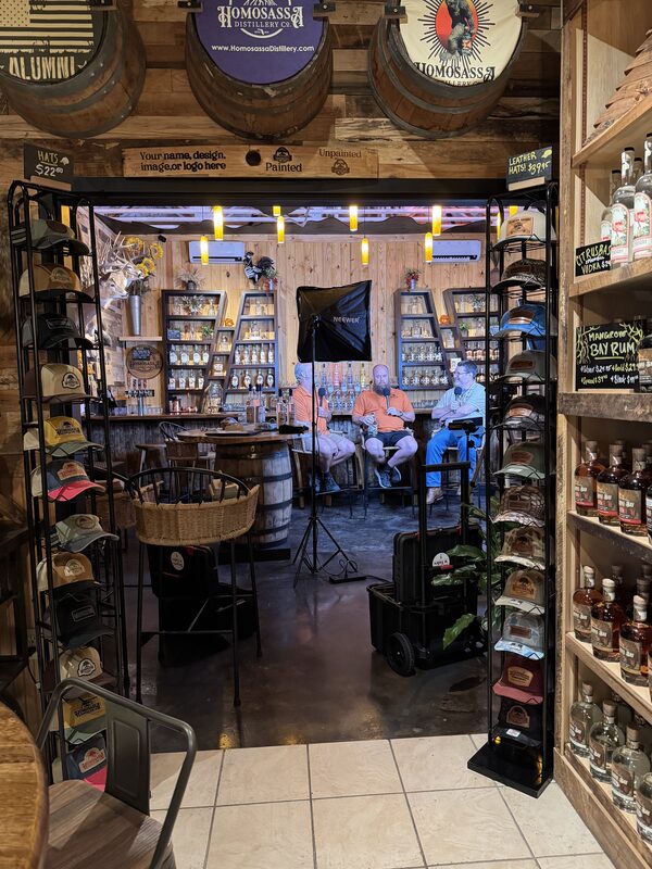 Interior view of a rustic distillery tasting room framed by hat racks and shelves of bottles, with wooden barrels and signage overhead. Three people sit on stools at the back behind a softbox light and camera equipment, surrounded by shelves of spirits and display items.