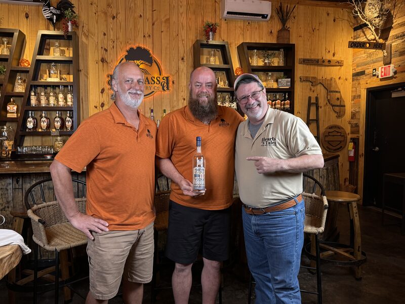 Three adult men stand in a wood-paneled tasting room; the center man holds a clear bottle of vodka while the two others stand on either side smiling. Shelves with bottles, a logo sign, wicker chairs and rustic decor fill the background.
