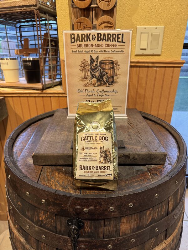 A wooden barrel serves as a display table holding a gold bag of "Cattle Dog" coffee labeled Bark & Barrel in front of a standing sign that reads "Bark & Barrel Bourbon-Aged Coffee" with an illustration of a dog and a barrel. The scene is indoors against wood-paneled walls with a light switch and a window shelf holding cups visible in the background.