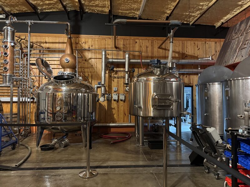 A room containing stainless-steel distillation and brewing equipment, including two large polished cylindrical tanks and a pot still with a copper swan-neck, connected by multiple pipes, valves and gauges mounted against a wood-paneled wall. The concrete floor has a central drain grate, hoses and tools scattered around, and an exposed insulated ceiling with additional metal tanks visible at right.
