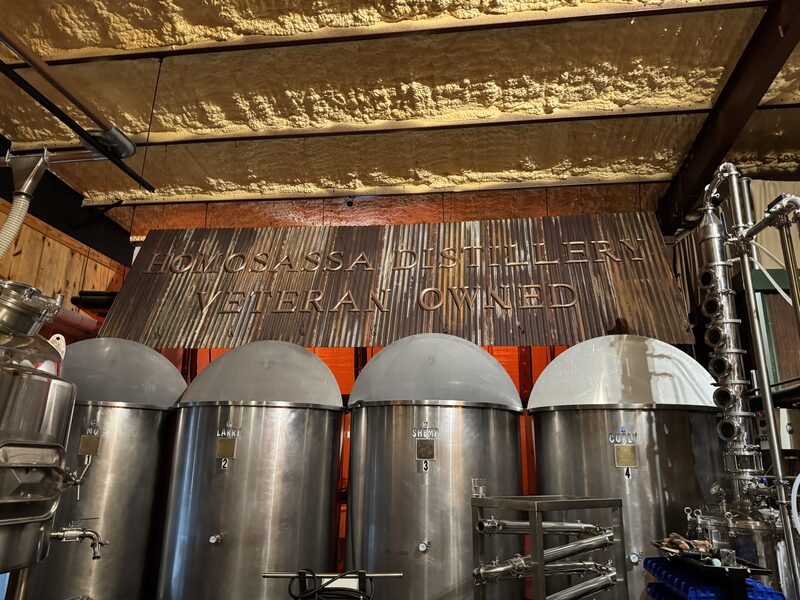 Four large stainless-steel fermentation tanks with rounded dome lids are lined up beneath a corrugated metal sign reading "HOMOSASSA DISTILLERY VETERAN OWNED." The industrial interior shows exposed insulated ceiling panels, pipes, and a vertical columnar still on the right.