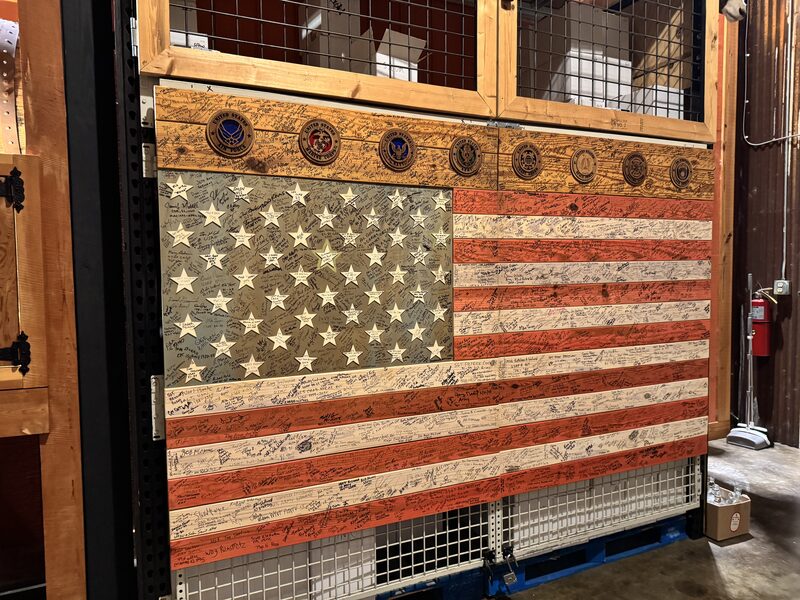 A large wooden American flag made from painted planks and cut-out stars is mounted on a metal grid and covered with numerous handwritten signatures across the blue field and red and white stripes. A row of circular military emblems runs along the top of the flag, and the scene shows a warehouse-like interior with a red fire extinguisher visible on the right.