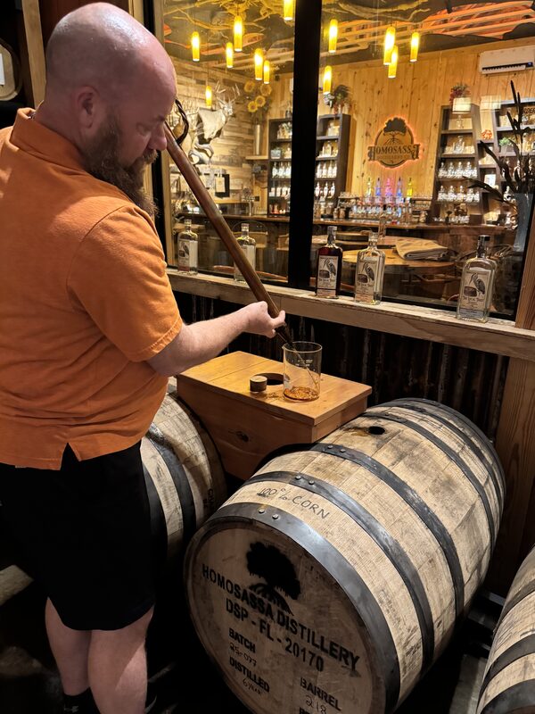 A bald, bearded man in an orange shirt uses a long wooden rod to draw amber liquid from a barrel into a glass set on a small wooden table. Several wooden barrels labeled "Homosassa Distillery" occupy the foreground and shelves of bottles, wooden paneling, and pendant lights are visible through a windowed bar area behind him.
