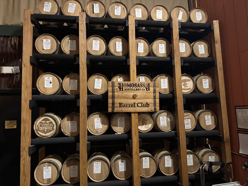 A wooden rack with five vertical columns of small oak barrels, each bearing a white tag, fills the frame. A wooden sign mounted near the center reads "Homosassa Distillery Co. Barrel Club" and a lower-left barrel shows a "Cypress & Matthews" logo.