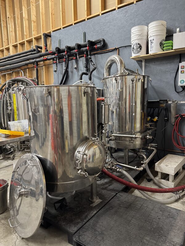 Multiple polished stainless steel brewing tanks and vessels with clamps, valves, and attached red and white hoses sit on a black rubberized floor mat. Behind them are exposed wooden studs and a gray wall with black insulated pipes, a small shelf holding white buckets, and a wall-mounted control panel.