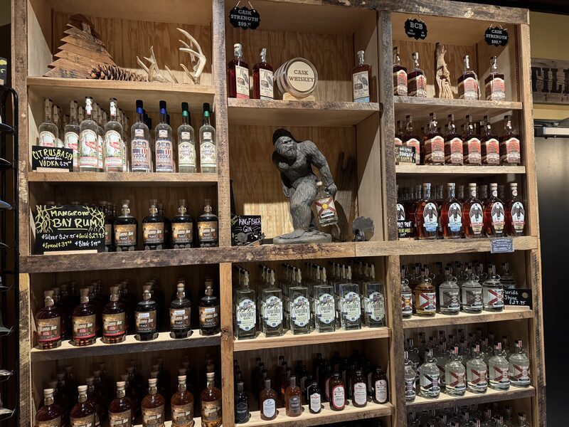 A rustic wooden shelving unit displays numerous bottles of liquor arranged in rows, small chalkboard price signs, antler decorations and a large gorilla statue holding a bottle in the central cubby. The bottles include varieties labeled vodka, rum and whiskey, with multiple identical bottles grouped together.