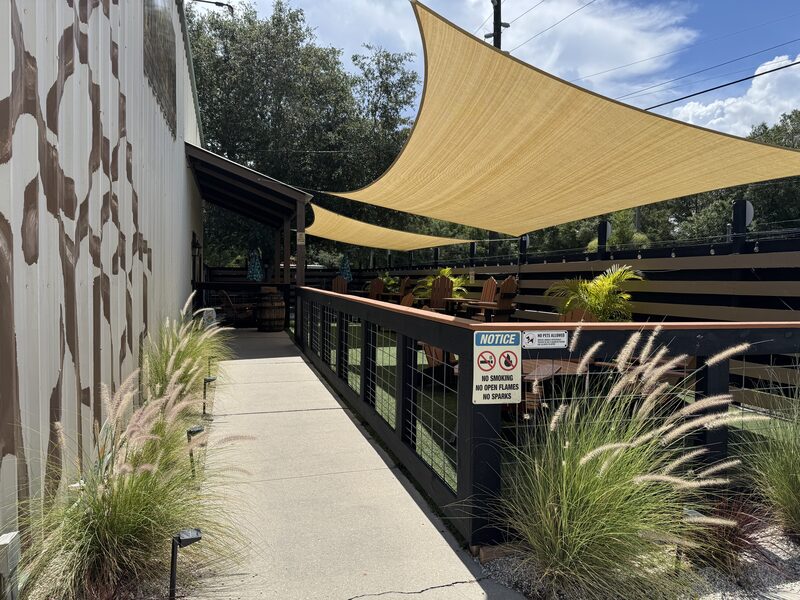 A sunlit outdoor patio area shows a concrete walkway bordered by ornamental grasses on the left and a fenced seating deck on the right with wooden Adirondack chairs under large beige triangular shade sails. A notice sign on the fence reads "NO SMOKING NO OPEN FLAMES NO SPARKS," and the scene includes a patterned metal wall, potted plants, and overhead power lines.