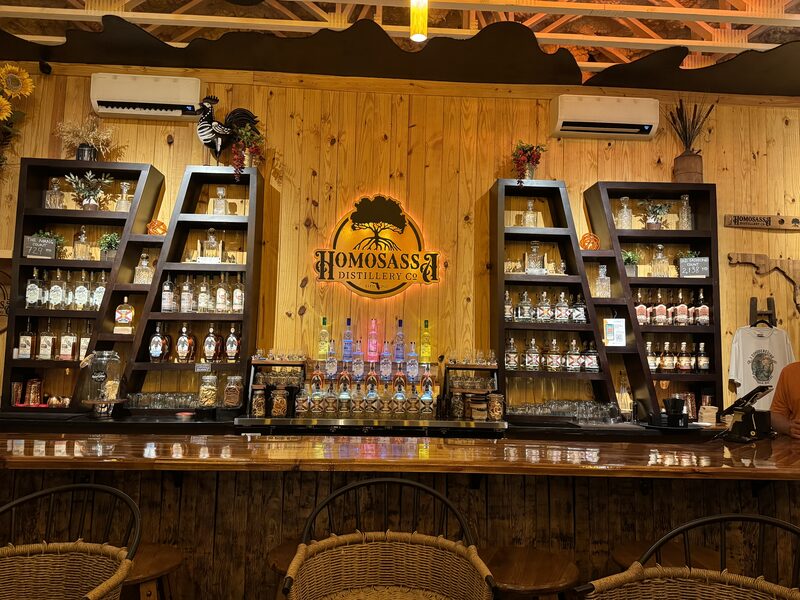 A warmly lit wooden bar interior features a central illuminated "Homosassa Distillery Co" sign flanked by two angled black shelving units filled with bottles, jars, and glassware, with a row of illuminated spirit bottles and taps on the countertop. Wicker bar stools sit in front of the polished wooden bar and decorative items like a rooster figure, potted plants, and wall-mounted air conditioners are visible above the shelves.