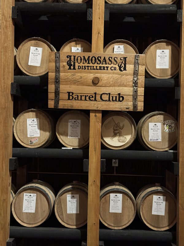 A wooden rack holding multiple small oak barrels with white labels, centered by a wooden sign reading "Homosassa Distillery Co. Barrel Club."