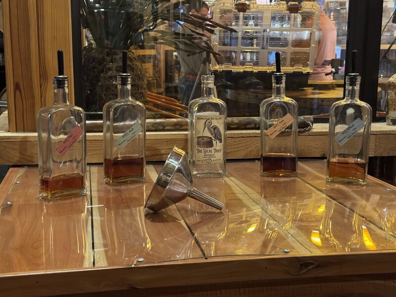 Five rectangular glass bottles with pour spouts and varying small amounts of amber liquid sit on a wooden counter, the center bottle bearing a white label with a bird illustration. A metal funnel lies on its side on the counter and a mirror behind reflects shelving and indistinct figures.