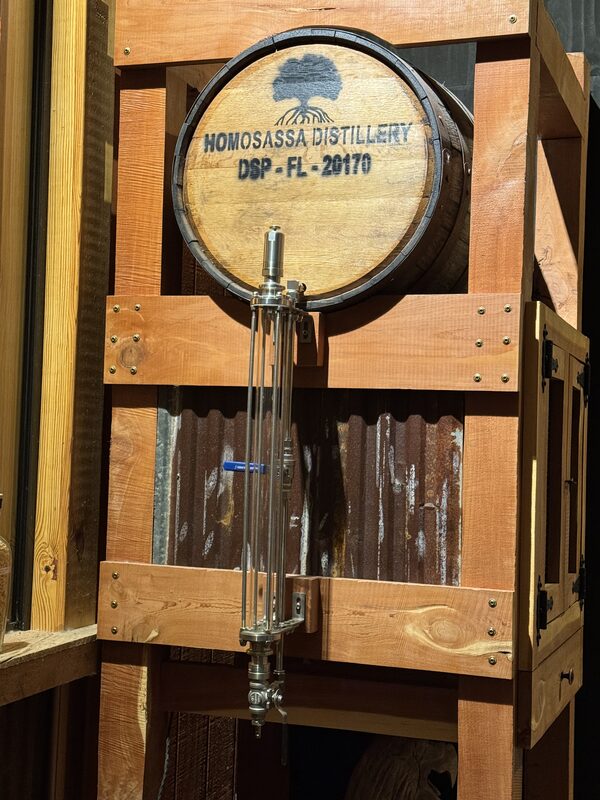 A wooden barrel stamped with a tree logo and the text "HOMOSASSA DISTILLERY DSP - FL - 20170" is mounted in a wooden frame above a vertical glass column and metal spigot assembly against a corrugated metal backdrop.