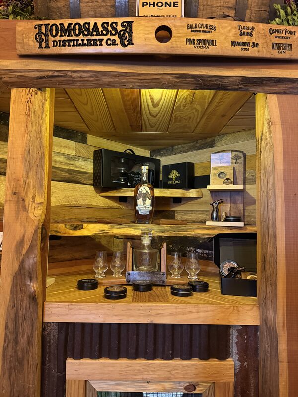 A rustic wooden display unit labeled "Homosassa Distillery Co." at the top contains a central bottle of liquor on the middle shelf. Surrounding it are several small tasting glasses, black lids or coasters, boxed packaging, a metal pourer or opener, and a wood-paneled backing lit with warm yellow lighting.