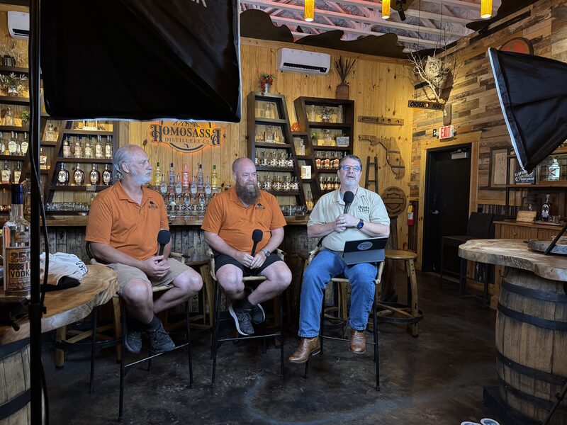 Three men sit on tall stools in a wooden distillery tasting room, two wearing matching orange shirts and the third in a light-colored shirt holding a microphone and a laptop. Professional softbox lights frame the scene while shelves of liquor bottles, wooden barrels, and rustic decor fill the background.