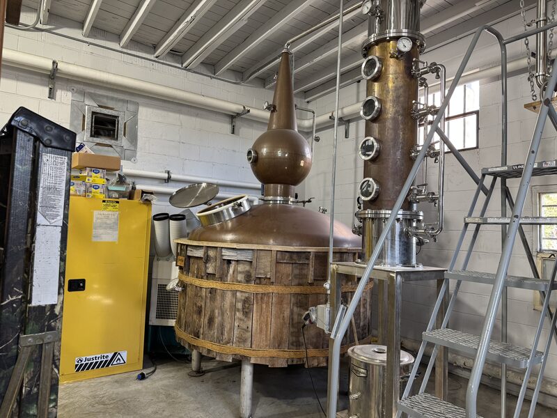 A distillery interior features a large copper pot still with a bulbous swan-neck head and a wooden staved outer jacket connected to a tall stainless-steel column with multiple circular sight glasses. A metal step ladder, a yellow cabinet, ducts, pipes, and exposed ceiling joists are visible around the equipment.