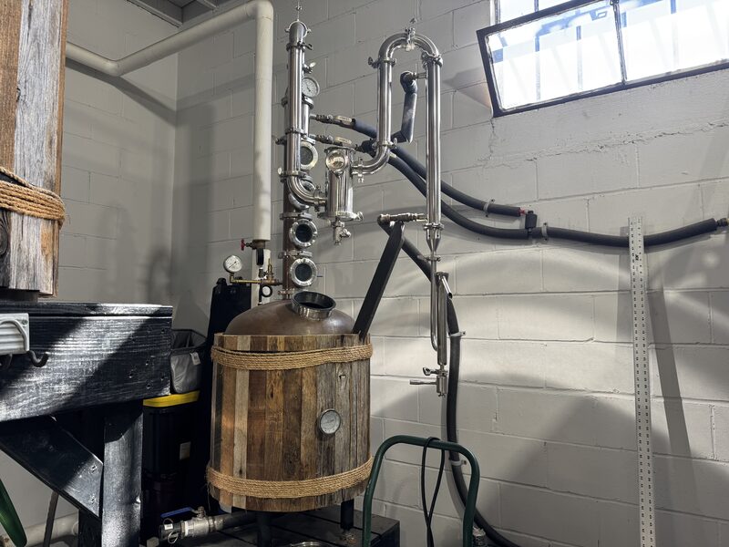 A wood-clad cylindrical metal vessel with a copper-domed top and an attached stainless-steel column featuring multiple sight-glasses, valves and pressure gauges stands against a white cinderblock wall, with black hoses connecting it to wall piping beneath a rectangular high window. Nearby are a dark wooden platform, a green hand truck, and a vertical metal ruler leaning against the wall.