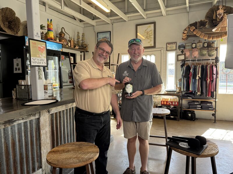 Two smiling middle-aged men stand in a rustic shop or tasting room; the man on the left points at a bottle the man on the right is holding. The space includes a corrugated metal bar with wooden stools, shelving and racks of hats and shirts, framed pictures on the walls, and sunlight streaming through windows.