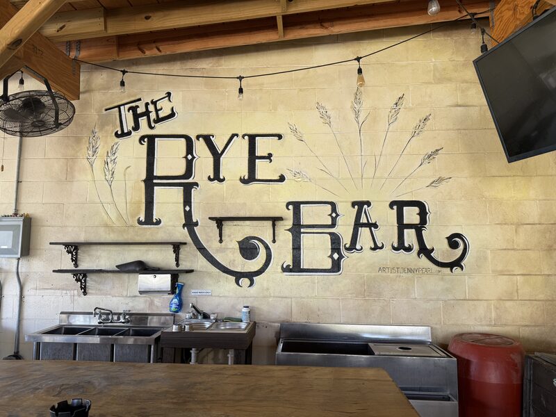 A rustic cinderblock wall features a large black hand-painted sign reading "THE RYE BAR" with painted wheat stalks, above metal sinks and counters, wooden shelves holding cleaning supplies, an overhead fan, and a mounted television.