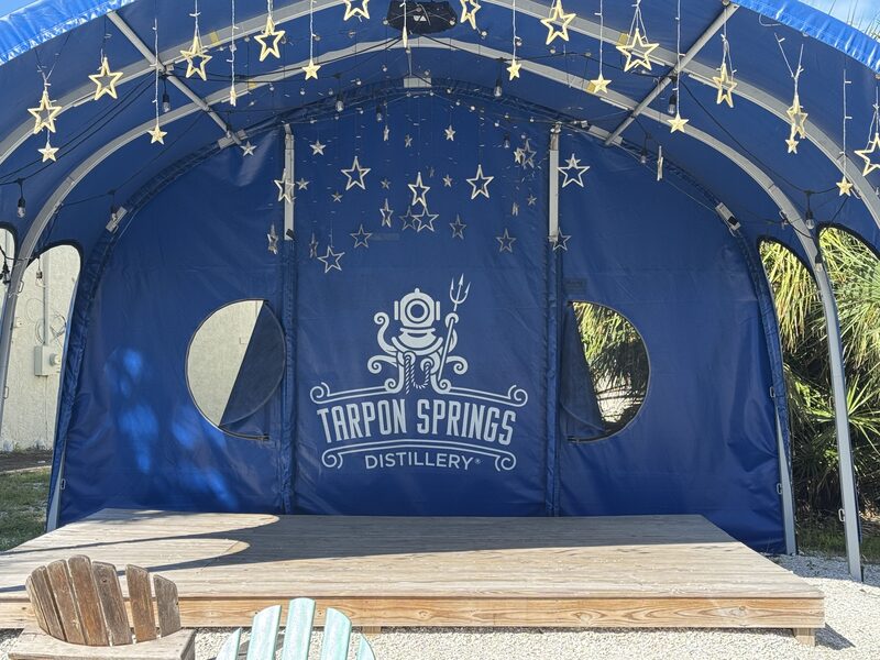 A blue fabric canopy/stage displays a large white "Tarpon Springs Distillery" logo centered on the back wall with two circular windows on either side and strings of star-shaped lights hanging from the frame. A wooden platform in front has a few Adirondack-style chairs at the edge and palm foliage is visible through the right circular window.