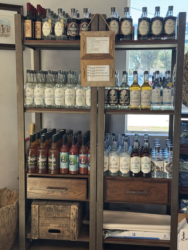 A wooden shelving unit holds multiple rows of assorted liquor bottles (clear, amber, and various labeled spirits) arranged across four main shelves with two wooden drawers and a wooden crate at the bottom, and a small double-framed sign hanging from the top center; a window to the right shows trees outside.