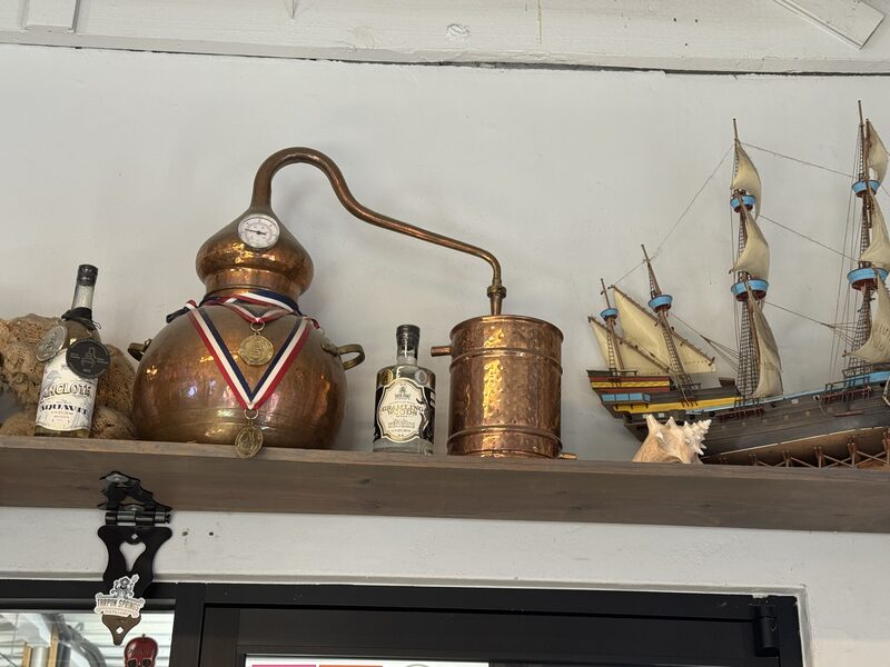 A high wooden shelf holds a copper alembic still with a pressure gauge and tricolor medals draped over it, flanked by two small glass bottles and a hammered copper container, with a model sailing ship and a conch shell to the right.