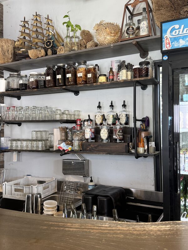 A café/bar service area with wall-mounted shelves holding rows of glass jars and bottles, multiple syrup pumps, stacked drinking glasses, utensils and small storage containers, plus decorative items like a model ship and a dried pufferfish on the top shelf. Below the shelves are a sink, dish rack, metal dispensers and a refrigerated display case at the right edge.