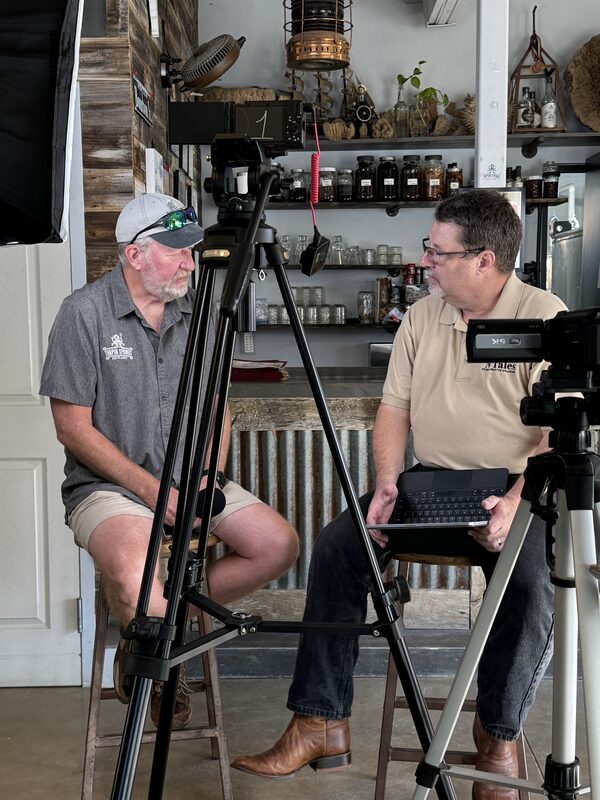 Two men sit on stools facing each other amid video production gear—two tripod-mounted cameras, a large softbox light, and a microphone—one holding a small laptop while the other wears a baseball cap with sunglasses perched on the brim. Behind them is a rustic counter with corrugated metal facing and shelves filled with glass jars, bottles, plants, and decorative items under a hanging lantern.