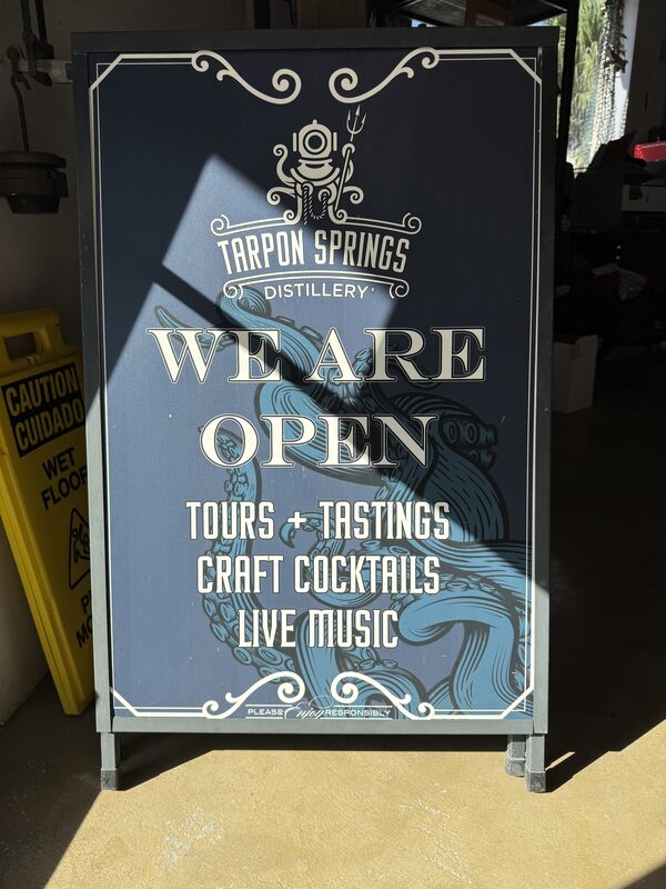 A freestanding dark blue A-frame sign with a decorative border and diver-helmet-and-tentacle illustration reads "Tarpon Springs Distillery" at the top and "WE ARE OPEN" in large letters, followed by "TOURS + TASTINGS," "CRAFT COCKTAILS," and "LIVE MUSIC." The sign stands on a concrete floor in a dimly lit interior with a yellow "Caution/Cuidado Wet Floor" sign to the left and shelving and boxes visible in the background.