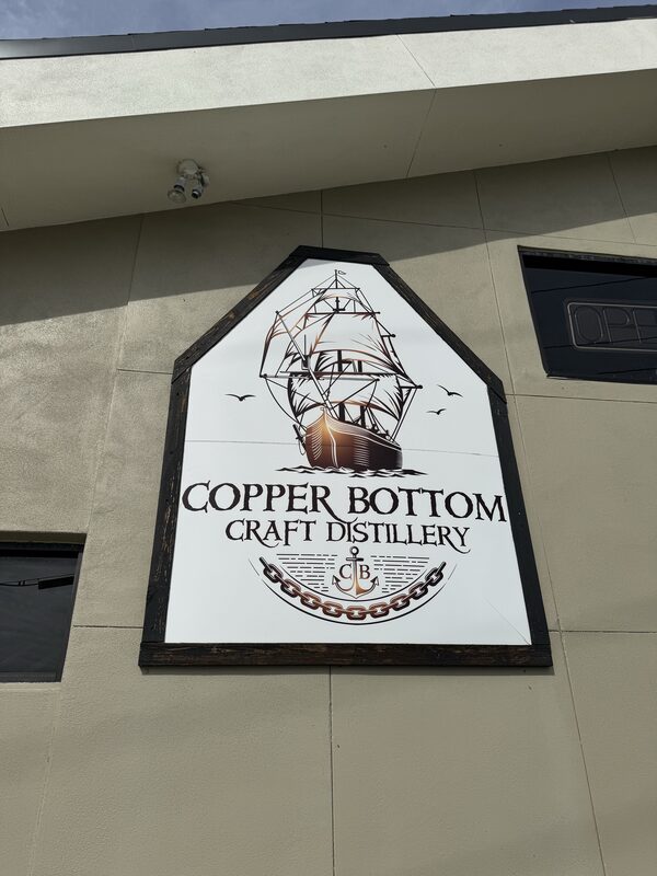 A large white, shield-shaped sign mounted on a beige exterior wall under an overhang features a stylized sailing ship with seagulls. Below the ship the text reads 'COPPER BOTTOM CRAFT DISTILLERY' above an anchor-and-chain logo.