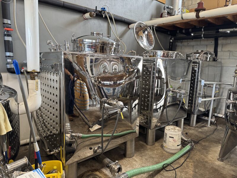 Several polished stainless-steel fermentation tanks with attached hoses, valves, and fittings sit on a concrete floor in an industrial room with exposed pipes and shelving, their mirrored surfaces reflecting nearby equipment and a partially visible person.