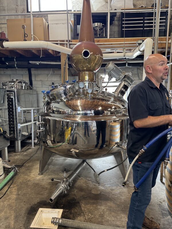 A large polished stainless-steel and copper still with gauges, valves, pipes and a tall conical copper column fills the center of an industrial room. A bald man in a dark short-sleeve shirt stands at the right holding blue hoses, with barrels, metal shelving and a concrete floor visible in the background.