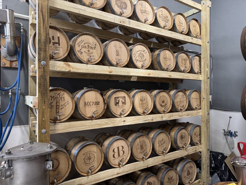 A multi-tiered wooden rack holding rows of small oak barrels with metal bands and spigots, each barrel face branded with different logos and labels. Blue hoses and stainless equipment are visible on the left, while a gray wall, plumbing fixtures, and a black trash bag appear on the right.