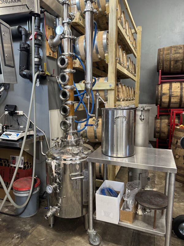 A stainless-steel pot still with a vertical column containing five circular sight-glass ports, a pressure gauge, and blue hoses stands beside a metal table holding a large stainless stockpot and cardboard boxes, with multiple wooden barrels stacked on a rack in the background.