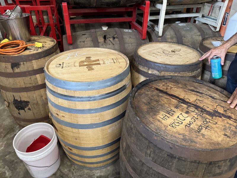 Several wooden barrels, including a newer light-colored barrel with metal hoops, are arranged on a concrete floor beneath red racks alongside an orange extension cord and a white plastic bucket containing a red cloth. A person's forearm and hand are visible at the right edge, resting on a barrel while holding a blue can.