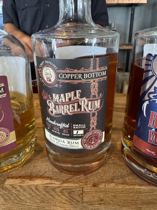 Close-up of a glass bottle labeled 'Copper Bottom Maple Barrel Rum' with an ornate black-and-red label reading 'Maple Barrel Rum' and 'Florida Rum,' containing amber liquid and sitting on a wooden countertop; the bottle is flanked by two other bottles and a person in a dark shirt is partially visible in the background.