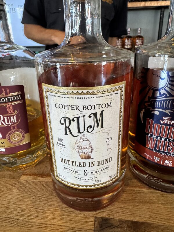 Close-up of a clear glass bottle containing amber liquid labeled 'Copper Bottom RUM' with a ship illustration and text 'Bottled in Bond,' '100 proof,' and '750 ml,' standing on a wooden surface between two other liquor bottles.