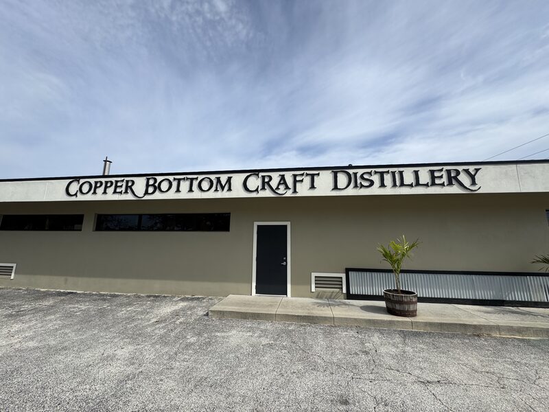 A single-story beige building displays large black letters across the top spelling 'COPPER BOTTOM CRAFT DISTILLERY,' with a centered dark door, long narrow windows on the left, and a potted palm in a wooden barrel to the right. A cracked asphalt parking area occupies the foreground and a wide, cloudy sky fills the background.