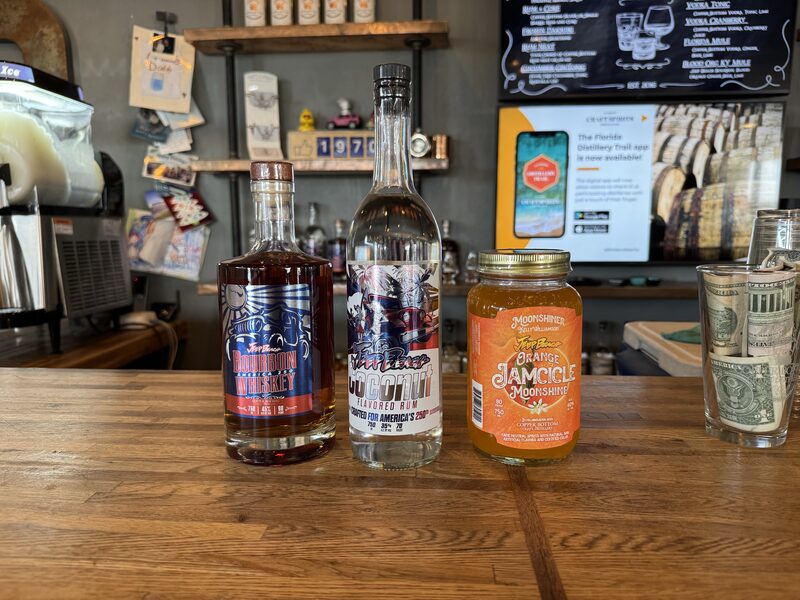 Three bottles sit on a wooden bar counter: a dark-brown Bourbon whiskey bottle at left, a clear coconut-flavored rum bottle in the center, and an orange &ldquo;Jamicile&rdquo; moonshine jar with a gold lid at right, with a glass of rolled US dollar bills beside them. Behind the counter are shelves with assorted items, a frozen drink machine at left, and a TV/menu screen on the wall.