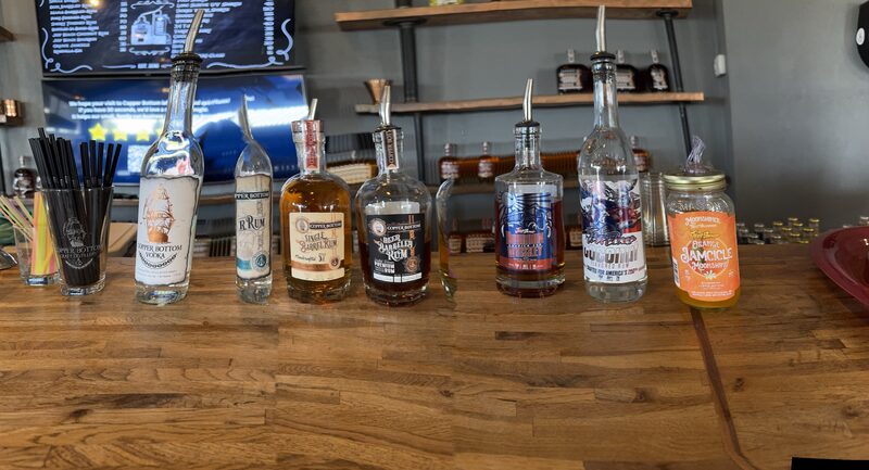A wooden bar counter displays a row of clear and amber liquor bottles with metal pour spouts, a glass of black straws on the left, and a mason jar with an orange label on the right. Shelves stocked with more bottles and a wall-mounted menu/TV are visible in the background.