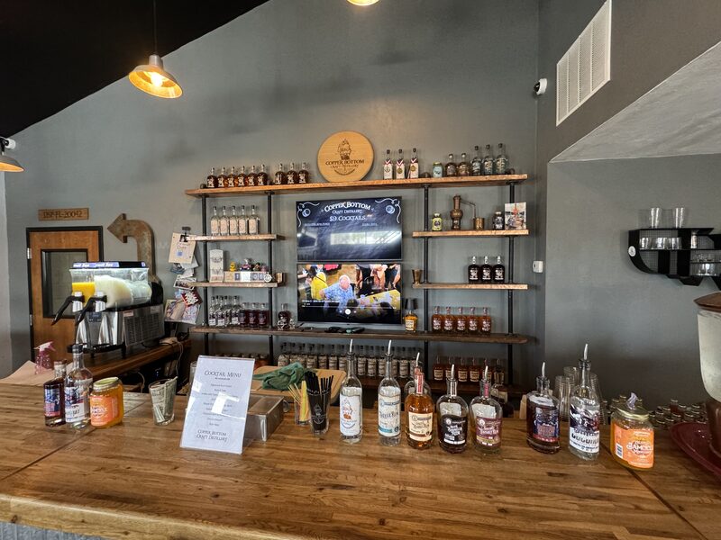 A wooden bar counter in the foreground displays assorted liquor bottles, jars, a container of straws, a printed cocktail menu, and a slushy machine to the left. Behind the counter is a wall of wooden shelves filled with more bottles and a central television screen showing people, with pendant lights hanging from the ceiling and gray painted walls.