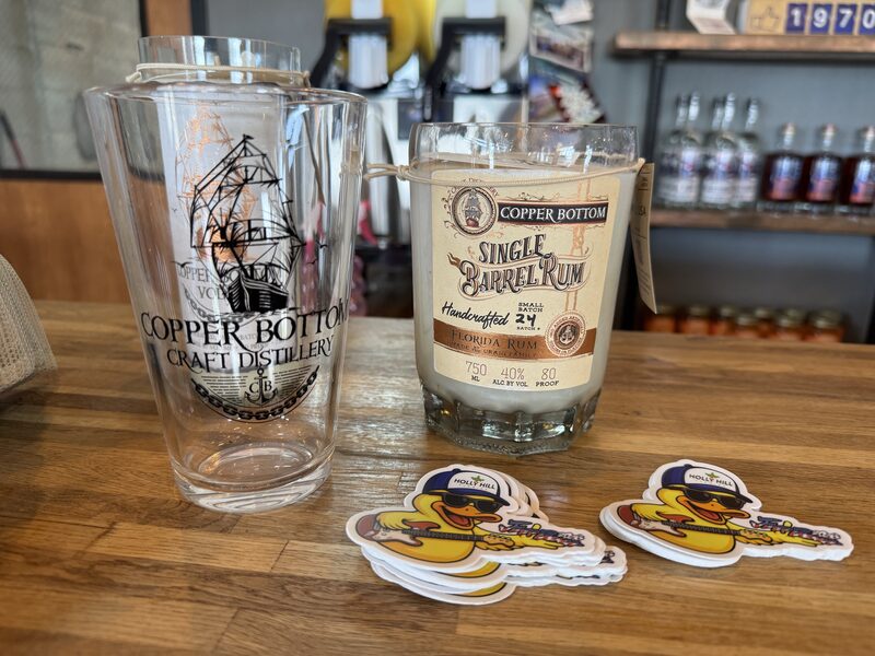 Two clear glass containers sit on a wooden counter: a tall pint glass on the left printed with a sailing-ship logo and the words 'Copper Bottom Craft Distillery,' and a shorter jar on the right labeled 'Copper Bottom Single Barrel Rum' with a hanging tag. In front of them are two small stacks of cartoon duck stickers wearing sunglasses and hats, with blurred shelves of bottles in the background.