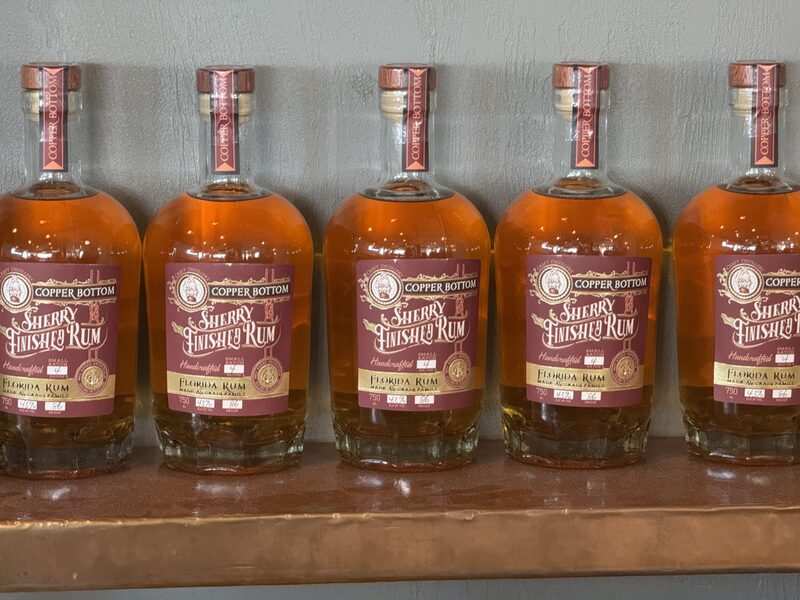 Five clear glass bottles of amber-colored rum with maroon 'Copper Bottom Sherry Finished Rum' labels and copper-colored neck seals are lined up on a wooden shelf against a gray wall.