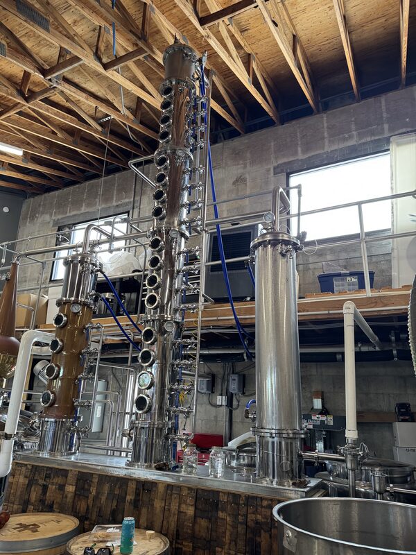 Interior of an industrial distillery showing tall stainless steel and copper column stills with multiple round sight-glass ports, connected piping, blue hoses and gauges mounted on a wooden-topped stainless workbench. Exposed wooden ceiling rafters, concrete block walls, a mezzanine with railings, barrels, a large metal tub and various tools are visible in the foreground.