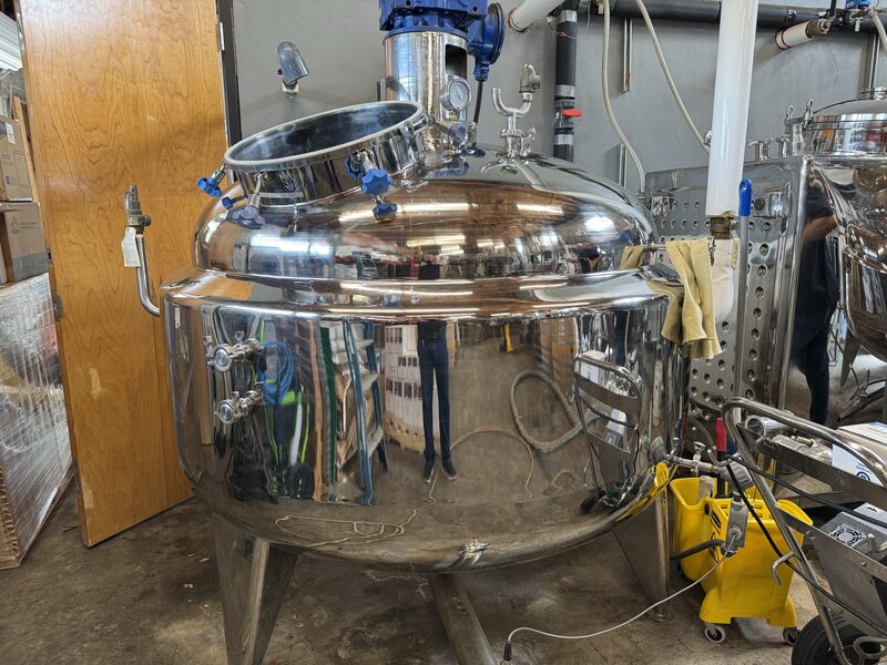 A large polished stainless-steel industrial tank with an open angled manway and blue-handled fittings stands on three legs in a workshop, its reflective surface showing distorted reflections of figures, shelving and equipment. Nearby are pipes, a hanging towel and a yellow mop bucket.